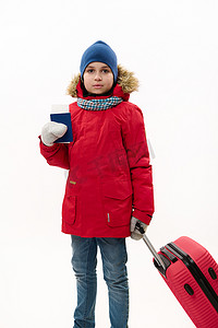 红色羽绒服摄影照片_穿着红色大衣和蓝色牛仔裤的十几岁旅行男孩带着登机牌和白色背景的手提箱走路