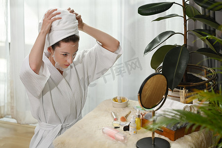 千禧一代女性头戴浴巾，敷在面罩上，看着梳妆台上的镜子