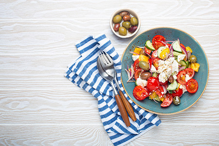 白色木质背景中蓝色碗中的希腊沙拉，配有菲塔奶酪、蔬菜和橄榄