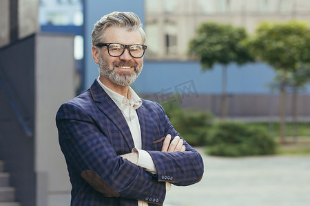 头发灰白的成熟投资者的画像，戴眼镜的老人看着相机和打手势，商务西装在办公楼外双臂交叉的商人，成功的老板银行家