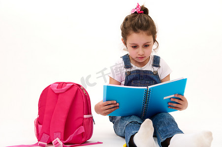 白人可爱的小女孩，一年级学生阅读教科书，坐在白色背景下的粉色背包旁