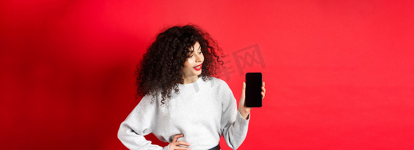 购物app摄影照片_一头卷发的时尚年轻女性的肖像显示空的智能手机屏幕，展示购物应用程序，站在红色背景上