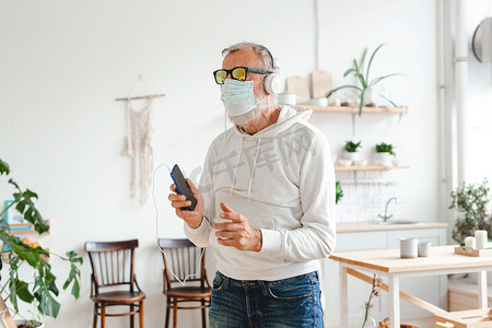 戴着医用面具的老人脸上跳舞，并使用智能手机应用程序在移动应用程序上创建播放列表音乐-老年男性用手机技术享受跳舞的乐趣-科技和快乐的老年生活方式概念