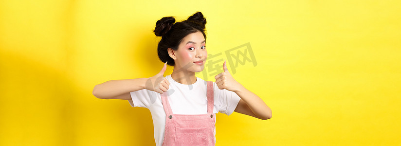 美丽的亚洲女孩，妆容亮粉色，竖起大拇指表示赞同，满意地微笑，推荐促销，黄色背景