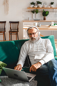 穿着连帽衫和眼镜的帅气老人正在使用笔记本电脑，一边坐在家里的沙发上一边微笑着听音乐