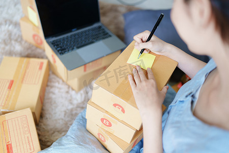 独立的年轻亚洲女性在线卖家的小企业主正在使用计算机并接受订单来包装产品以交付给客户。