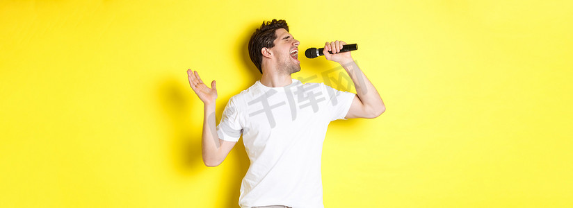 年轻男歌手拿着麦克风，唱高音，唱卡拉 OK，站在黄色背景上