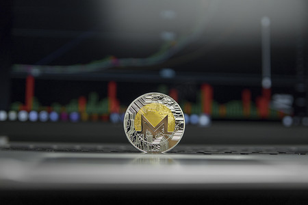 monero摄影照片_银色笔记本电脑黑色键盘上的 Monero 金币和银币，以及屏幕上的图表图表作为背景。
