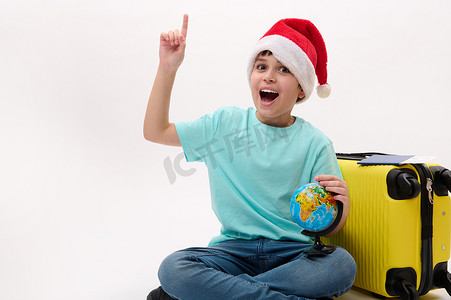 戴着圣诞帽和夏装的快乐青春期男孩，带着黄色手提箱和地球仪，指着白色背景上的复制空间