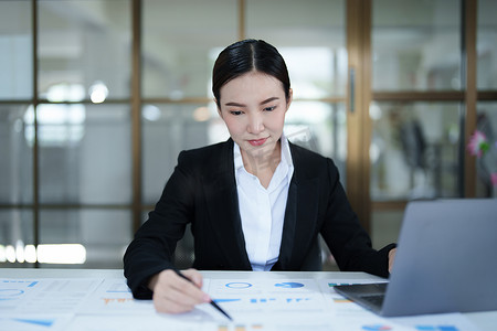 一位深思熟虑的亚洲女商人的肖像，她正在使用办公桌上的电脑查看财务报表并制定营销计划