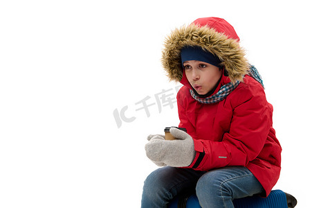 红色羽绒服摄影照片_穿着红色羽绒服和羊毛手套的可爱旅行男孩去度假，用一杯热茶暖手