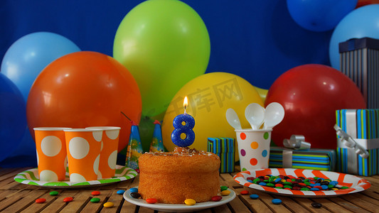 气球墙摄影照片_在土气木桌上的生日蛋糕有五颜六色的气球、礼物、塑料杯子和塑料盘子的背景有糖果和蓝色墙壁的在背景中