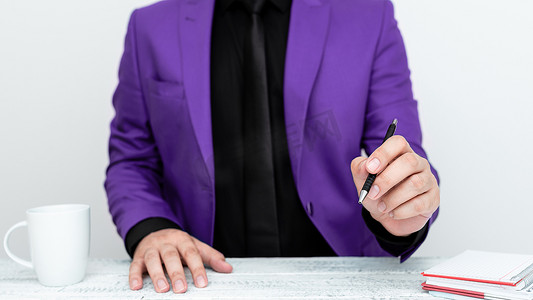 西装套装男摄影照片_穿着紫色西装的男模特坐在白桌旁，用笔指着重要信息。