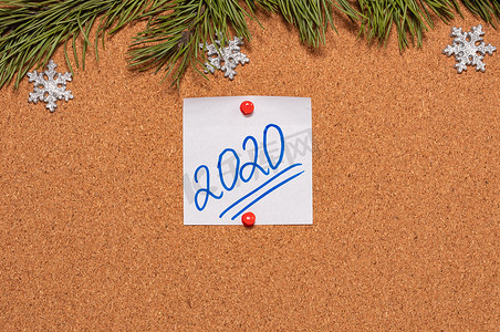 带有 2020 年手写体的白色便签贴在布告板上，布告板上装饰着松树枝和雪花。 