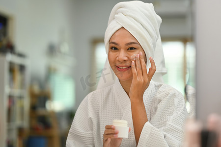头戴毛巾的微笑美女在脸上涂滋养霜，享受面部护肤程序