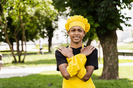 黑人厨师摄影照片_穿着厨师帽和黄色围裙制服的黑人非洲少年厨师在户外微笑。