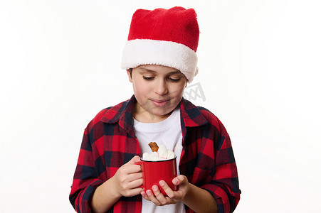 戴着圣诞帽的可爱的青春期前男孩，用一杯红色的热巧克力和棉花糖温暖他的手，与白色隔离