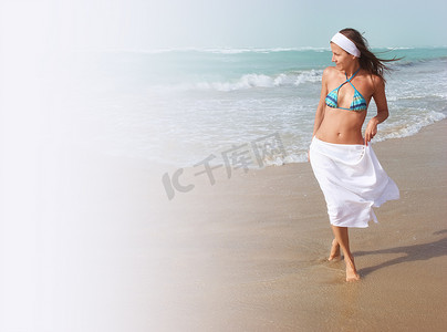 吹裙子摄影照片_美丽的年轻女人赤脚走在沙滩上，沿着海洋，风在吹，头发和白裙子