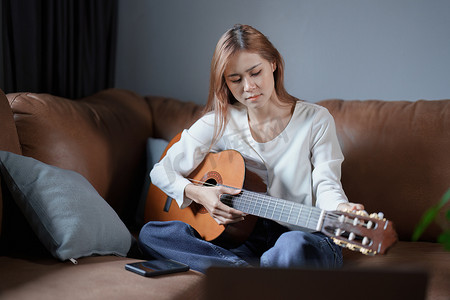 坐弹吉他摄影照片_亚洲年轻女性在沙发上弹吉他放松度假压力的画像