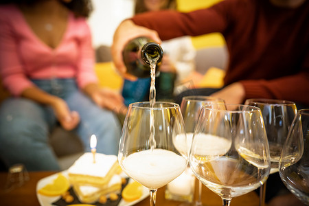 喜事摄影照片_将闪闪发光的白葡萄酒倒入高脚杯和其他玻璃杯和生日蛋糕中的特写 — 面目全非的年轻人在家里喝香槟庆祝喜事