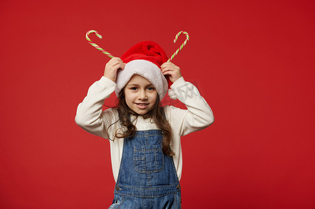 戴着圣诞帽的快乐小女孩头上拿着条纹棒棒糖，像喇叭一样，微笑着看着镜头，红色背景