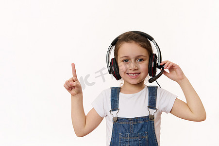 戴耳机的女生摄影照片_聪明的小女生，戴着耳机的一年级学生，可爱地微笑着指着白色背景中的复制空间