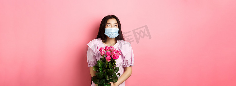 戴着医用面具的浪漫亚洲女孩用梦幻般的眼睛望着空荡荡的空间，在情人节拿着一束玫瑰，在大流行病期间与情人约会