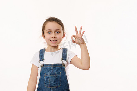 儿童展示摄影照片_可爱的小女孩，穿着蓝色牛仔工作服，手势，展示和平标志，对着镜头可爱地笑着欢快的露齿微笑