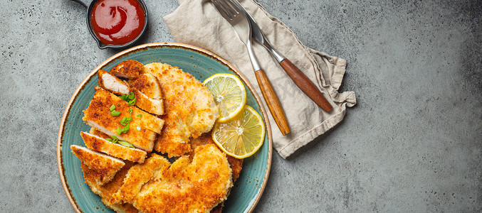 香脆的 panko 面包屑炸鸡片配绿色沙拉和柠檬切在灰色背景桌上的盘子上，上面放着番茄酱。