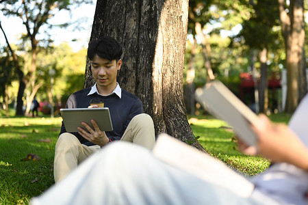 微笑的亚洲男学生使用平板电脑，在校园树下准备考试。