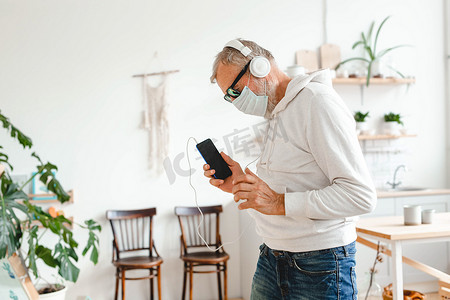 戴着医用面具的老人脸上跳舞，并使用智能手机应用程序在移动应用程序复制空间上创建播放列表音乐-老年男性享受与手机技术共舞的乐趣-科技和快乐的老年生活方式概念