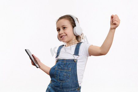 儿童智能摄影照片_带着智能手机的快乐小女孩，在立体声耳机上听音乐，在白色背景中被隔离，有复制空间