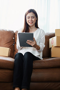 独立的年轻亚洲女性在线卖家的小企业主正在使用平板电脑并接受订单来包装产品以交付给客户。
