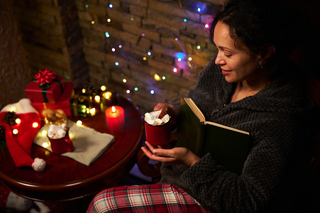 寒假阅读摄影照片_可爱的女人拿着一杯热巧克力，喜欢在烛光下阅读。
