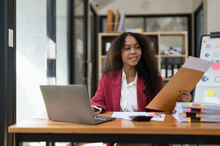 坐着的会计摄影照片_微笑的年轻非洲裔美国女性金融企业家坐在工作场所的办公室里，端着咖啡杯，在笔记本电脑上工作，投资财务文件报告财务分析概念。