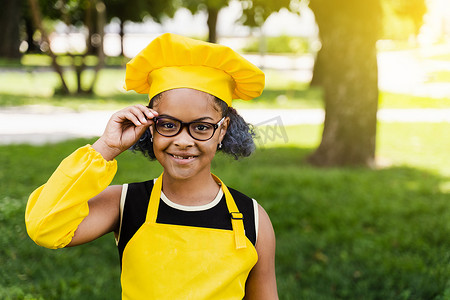 穿着厨师帽和黄色围裙制服的非洲黑人儿童厨师女孩摸着 hil 眼镜，在户外微笑。
