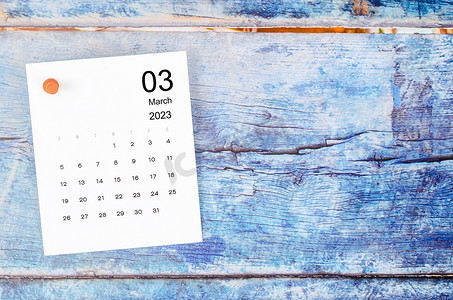 2023 年 3 月的月历和蓝色木质背景上的木制图钉。