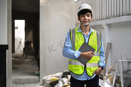 身穿安全头盔和反光背心的建筑师站在建筑工地前，对着镜头微笑的肖像