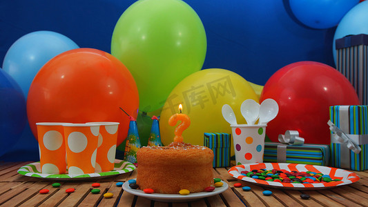 带蜡烛的生日蛋糕，在质朴的木桌上呈问号形状，背景是彩色气球、礼品、塑料杯和塑料盘，背景是糖果和蓝色墙壁