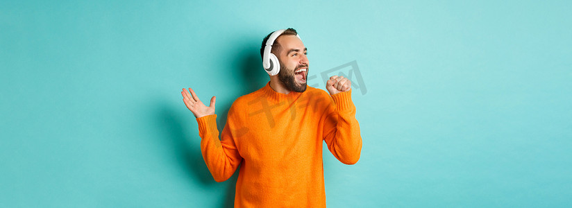 快乐的男人一边用耳机听音乐，一边拿着隐形麦克风，站在绿松石色的背景上同步口型