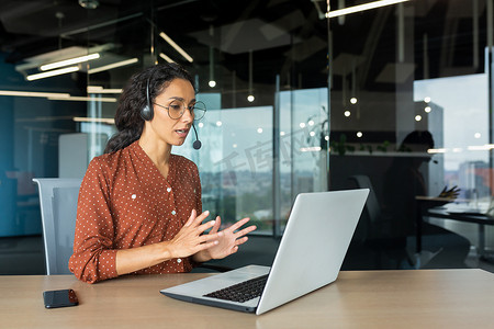 与同事进行视频通话在线会议，在现代办公室工作的西班牙裔女性，女商人微笑着使用笔记本电脑和耳机远程交谈