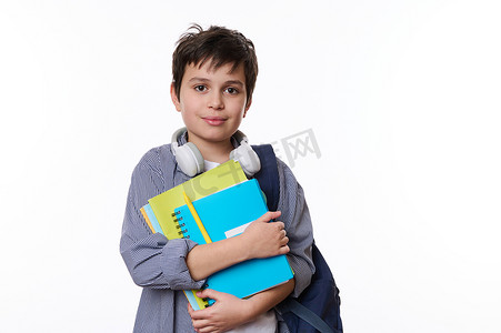 自信的少年戴着耳机，看着相机，在白色背景下摆着学校用品和背包