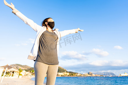 在撒丁岛 Golfo Aranci，戴着黑色医用口罩的年轻女性将双臂伸向蓝天，望着落日 — 尽管有冠状病毒，但冬天在海上的愉快生活方式