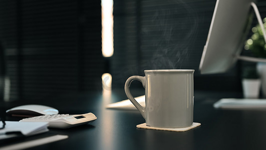 一杯热咖啡摄影照片_黑色木桌上的一杯热咖啡，背景是电脑、文档和计算器。