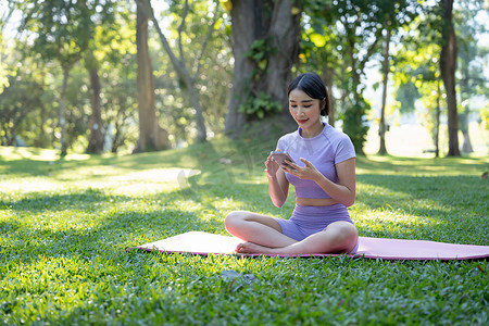 亚洲年轻女性在夏季公园练习时使用手机坐在瑜伽垫上