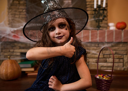 巫师摄影照片_特写哥特式小女孩，脸颊上画着蜘蛛，戴着巫师帽，竖起大拇指看着镜头