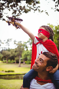 穿着超级英雄服装的女孩坐在父亲的肩膀上，拿着一架玩具飞机