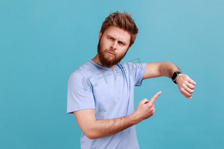 迟到早退摄影照片_穿蓝色 T 恤的男人用手指指着手表，不耐烦地检查时间是否迟到