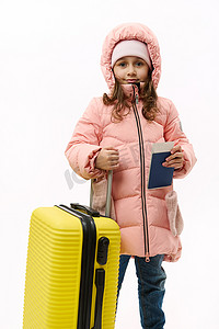 一个穿着粉色羽绒服的小女孩的全长肖像，在白色背景上摆着手提箱和登机牌