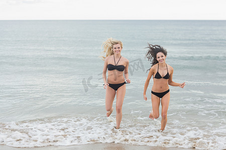 海滩奔跑的美摄影照片_微笑的比基尼女性在海滩的水中奔跑
